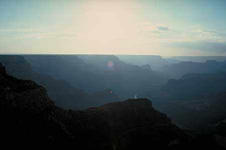 Silhouetted landscape of the Grand Canyon at dusk, with soft, fading sunlight creating a hazy, bluish atmosphere. The vast rock formations and a winding river are visible in the distance under a clear sky. The rock cliffs are less and less visible in the distance due to the scattered light. This is an illustration of what is called atmospheric perspective and how we use it as a clue to the perception of spatial depth.