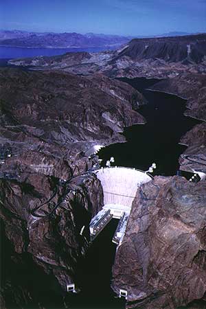 Aerial view of a Hoover Dam holding back Lake Mead, surrounded by rugged, brown mountains. Roads and structures are visible on the dam. The sky is clear, and distant mountains are visible in the background. It is an illustration of what is called bird´s eye perspective.
