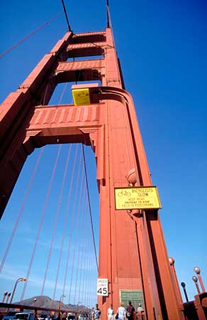 The image shows a tall, red tower of the Golden Gate Bridge against a clear blue sky. A speed limit sign of 45 mph is visible near the bottom. People are walking on the pathway at the base of the tower. As the tower is shown from the bottom upwarts, it is an illustration of what is called frog perspective.