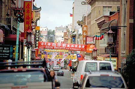 A busy street in San Franciscos Chinatown with various banners and signs in Chinese. Cars line both sides of the road. Decorative lanterns hang above, and buildings on either side display traditional architecture. It is an illustration of what is called eye perspective.
