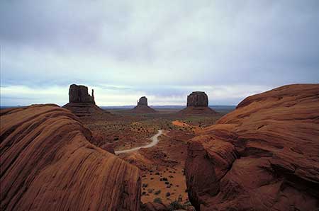 A vast desert landscape with three towering rock formations under a cloudy sky. The foreground is framed by rugged brown cliffs, and a winding dirt road crosses the arid terrain. The scene is Monument Valley and it illustrates how we use the overlapping of objects as a clue to perceive spatial depth.