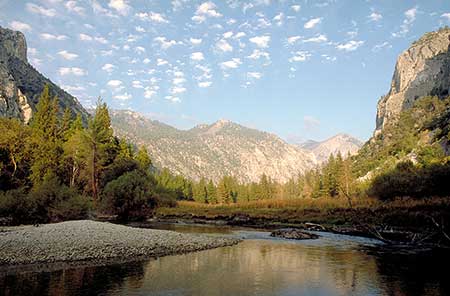A serene landscape with a tranquil river flowing through a valley surrounded by tall pine trees and rocky cliffs under a sky dotted with clouds. It is a wide-angle shot and so the trees in the foreground are imaged much taller than those in the background. This is an illustration of how we use relative size of objects to construct spatial depth.