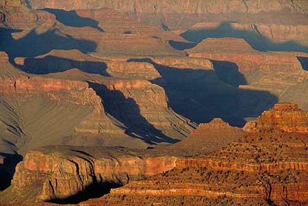 A scenic view of the Grand Canyon at sunset, showcasing its vast layers and formations. The warm sun casts dramatic shadows over the rocky ridges, highlighting the diverse textures and deep, rich colors of red, orange, and brown. It is an illustration about how our visual perception uses cast shadows to construct spatial depth.