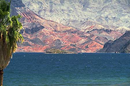 Desert landscape featuring the blue water of a lake in the foreground and rugged red rock formations in the background, with a mountain range in the background under a clear blue sky. Sparse vegetation is visible across the rocky terrain. It is an example of an image that uses the color perspective to create the peception of spatial depth.