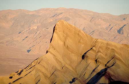 A rugged, sunlit peak rises sharply amid a desert landscape, with layers of barren mountains in the background under a clear sky. It depicts Manley Beacon in Death Valley. It is an example of an image with little spatial depth effect due to weak color contrast.