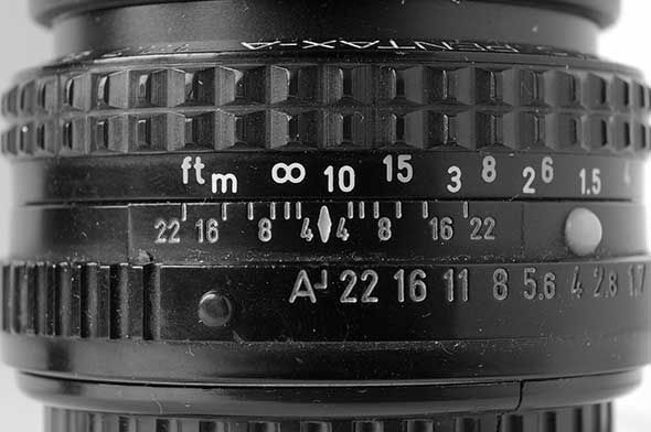 Close-up of a camera lens with focus and aperture settings visible. The focus scale shows distances in feet and meters, and the aperture scale displays f-stops ranging from 22 to 2.8. The lens is predominantly black with textured grips.
