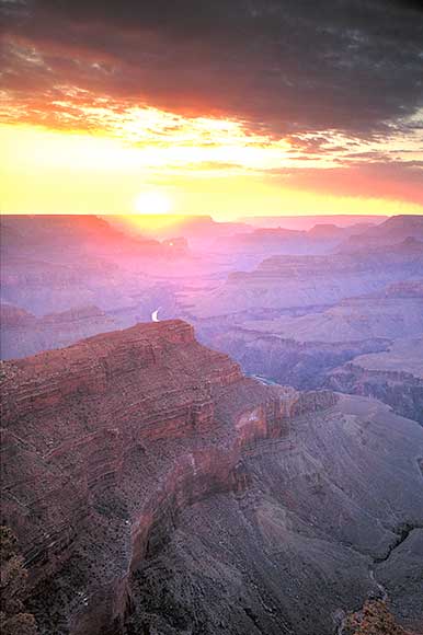 Sunset over the Grand Canyon, with the sun partially hidden by clouds. The sky is a blend of orange, pink, and purple hues, casting a warm glow on the canyons layered cliffs and deep valleys. The foreground is sharply focused.