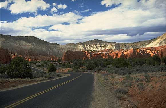 A landscape view of a desert with vibrant red and orange rock formations under a clear blue sky. Sparse vegetation is scattered across the foreground. Hills and mountains can be seen in the background.