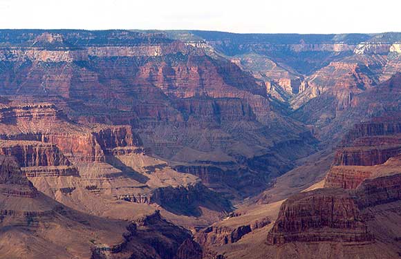 View of the Grand Canyon with layered rock formations, steep cliffs, and deep valleys. The hues of red, brown, and orange are prominent, showcasing the natural beauty of the landscape under a clear sky.