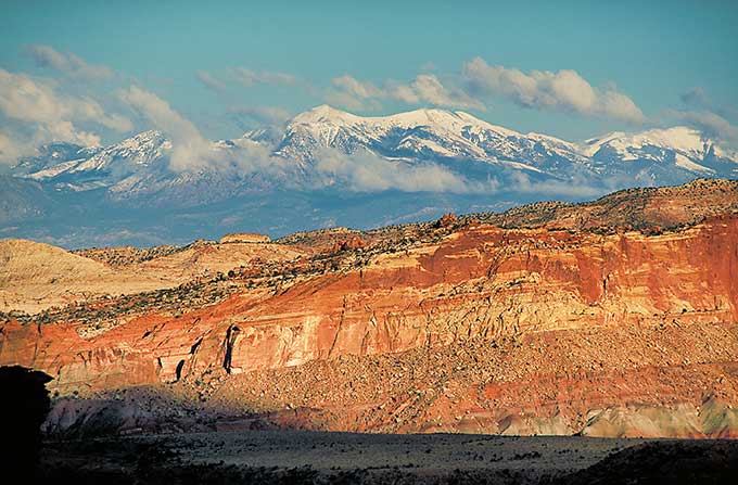 An image of the rugged form of he waterpocket Fold in Capitol Reef NP, glowing red in the afternoon light, with some snowcovered mountains in the distance.